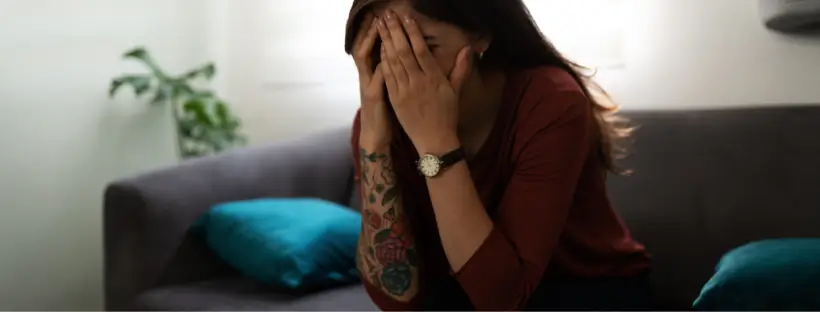 A woman with tattoos wearing a brown shirt and black watch sitting on a couch, covering her face with her hands, depicting stress and overwhelm, symbolizing early signs of hormonal imbalance.
