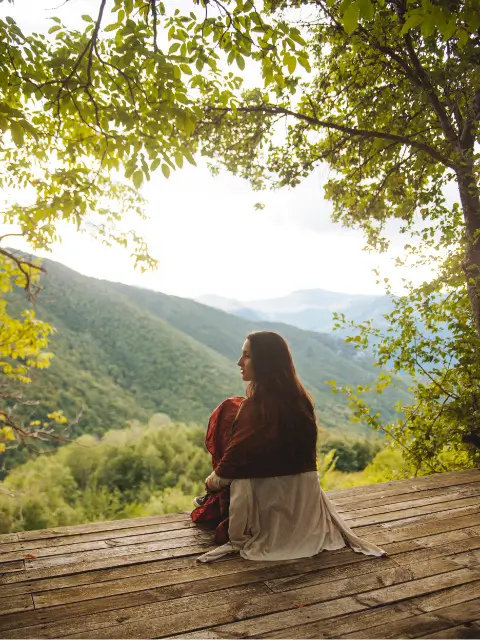 holistic-wellness-mountain-retreat-mindful-living Woman sitting on a wooden deck overlooking forested mountains, reflecting in a calm natural setting – symbolizing holistic wellness and mindful living.