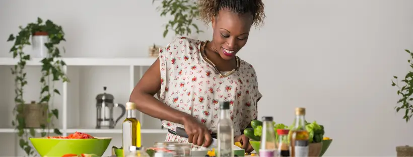 Woman smiling and preparing fresh vegetables in a bright kitchen, surrounded by bottles of oil and green plants.